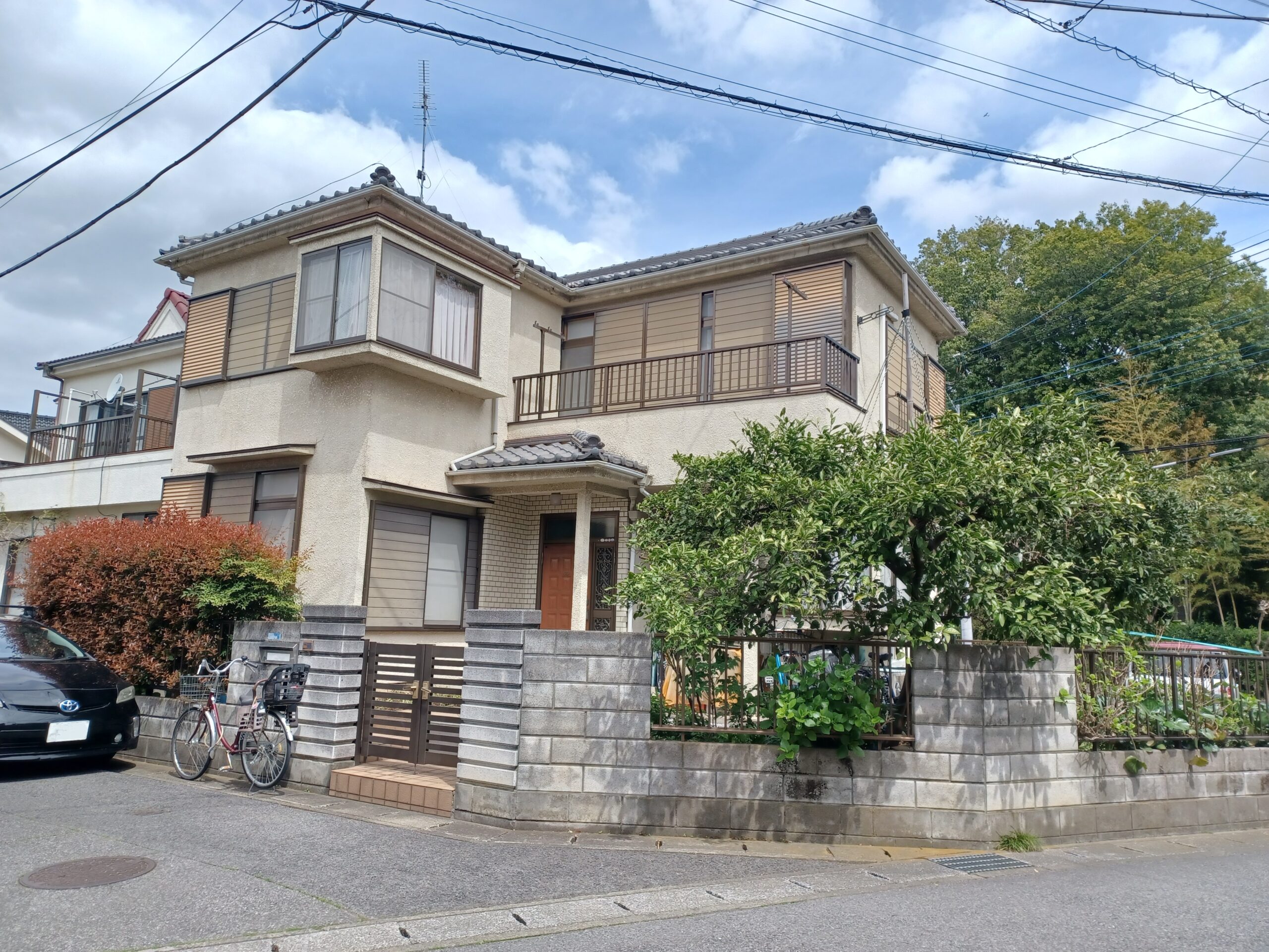 外壁塗装工事の施工前の写真、家全体を引いてとっている。2階建ての家で白い外装、家のまわりは低い塀で囲んであり、写真中央やや左に低い門扉がある